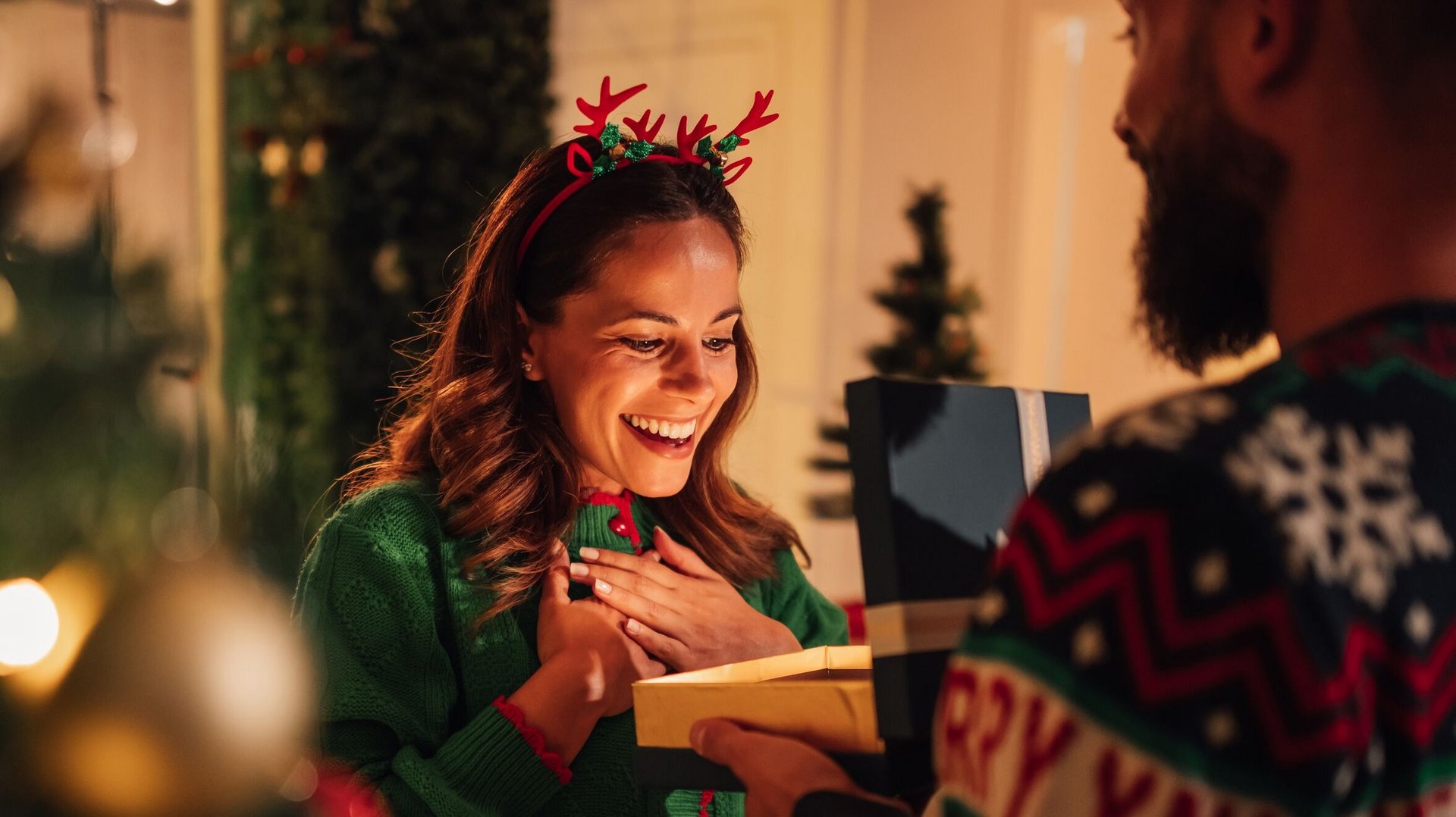Frau in Weihnachtspulli erhält von einen Mann ein Geschenk und freut sich