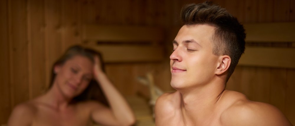 Junger Mann sitzt in der Sauna, Frau mit weißem Handtuch im Hintergrund